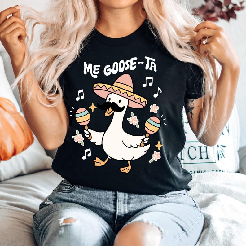 May include: Black t-shirt with the text "Me Goose-Ta" and a cartoon goose wearing a sombrero and mustache, holding maracas. Musical notes and floral accents surround the goose, creating a festive design.