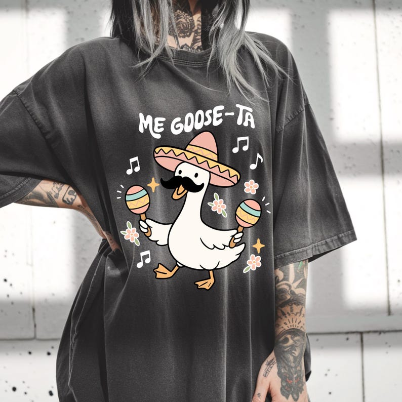 May include: Dark gray t-shirt featuring a cartoon goose wearing a sombrero and a mustache, holding maracas. The text "ME GOOSE-TA" is above the goose, with musical notes and floral accents around the design.