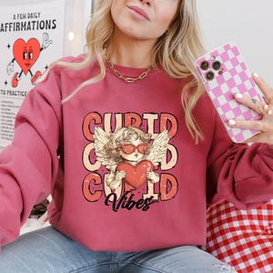May include: A pink sweatshirt with a graphic of a cupid holding a heart and the text "Cupid Vibes".