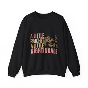 May include: Black sweatshirt with the text "A Little Ratchet A Little Nightingale" in pink and beige. A graphic of a genie lamp and skeletal hands is on the right side of the text.