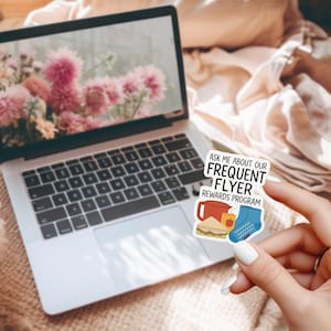 May include: A laptop displays a floral wallpaper. A sticker, held in front of the laptop, reads "ASK ME ABOUT OUR FREQUENT FLYER REWARDS PROGRAM". The sticker features illustrations of a mug, sandwich, apple, and a sock.