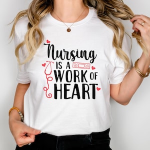 Op de afbeelding: Wit T-shirt met een zwarte afbeelding waarop staat "Nursing is a work of heart". De afbeelding bevat een rode stethoscoop en een rood pleister.