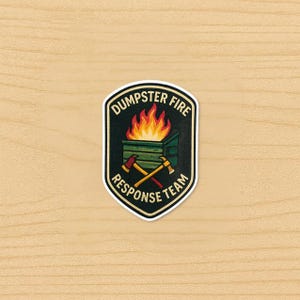 Peut inclure: Un autocollant avec une bordure noire et les mots "DUMPSTER FIRE RESPONSE TEAM". L'autocollant représente une benne à ordures avec des flammes et des haches croisées. Le fond est un grain de bois clair.
