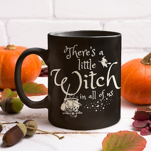 May include: Black coffee mug with a white design that says "There's a little Witch in all of us". The design includes a witch flying on a broomstick and a cauldron with a wand. The mug is surrounded by pumpkins and fall leaves.