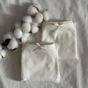 Può includere: Due paia di biancheria intima in pizzo bianco con un motivo delicato. La biancheria intima ha un fiocco sul davanti e un bordo in pizzo.