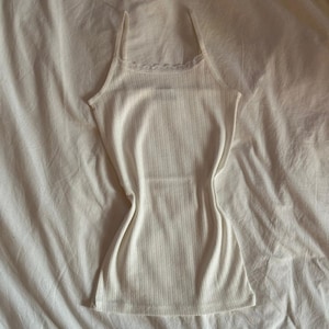 May include: A white ribbed tank top with a lace trim around the neckline. The tank top is laying flat on a white bedspread.