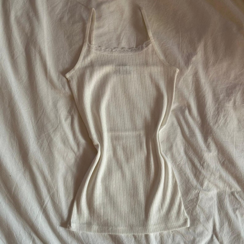 May include: A white ribbed tank top with a lace trim around the neckline. The tank top is laying flat on a white bedspread.