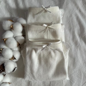 May include: Three pairs of white cotton underwear with lace trim and a bow detail. The underwear is stacked on top of each other.