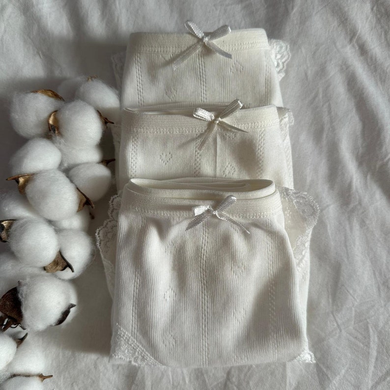 May include: Three pairs of white cotton underwear with lace trim and a bow detail. The underwear is stacked on top of each other.
