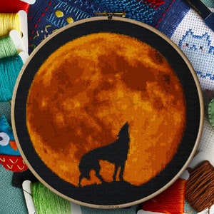 Könnte beinhalten: Kreuzstich-Stickrahmen mit einer schwarzen Silhouette eines heulenden Wolfs vor einem großen orangefarbenen Mond. Der Rahmen ist von bunten Stickgarnen, einem Knopf und kleinen Filzarbeiten umgeben.