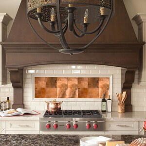 May include: A kitchen with white subway tile backsplash, a copper tile accent wall, and a black chandelier over a gas stove. The countertop is a dark brown granite with a wooden chopping board and a loaf of bread on it.