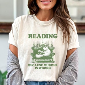 May include: A white t-shirt with a green graphic of a cat reading a book in a chair. The text on the shirt reads "Reading because murder is wrong."