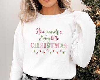May include: White sweatshirt with the text "Have yourself a Merry little CHRISTMAS" in pink and green script. The word CHRISTMAS is in a block letter style with pink and green accents. A string of colorful Christmas lights is below the text.
