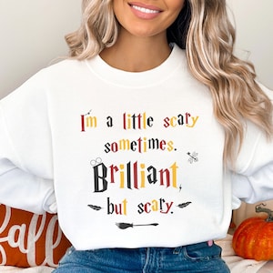 May include: White sweatshirt with the text "I'm a little scary sometimes. Brilliant, but scary." in various colors. The design includes a broom, glasses, and a lightning bolt. A fall-themed pumpkin is in the background.