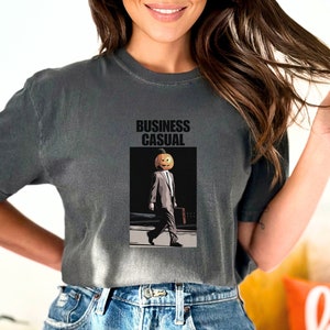 May include: Dark gray t-shirt with the words "BUSINESS CASUAL" above an illustration of a person with a pumpkin head wearing a suit and carrying a briefcase. The person is walking on a crosswalk.