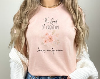 May include: A light pink t-shirt with the text "The God of Creation knows me by name" in black script. A floral design in shades of pink and peach is centered above the text. The shirt is made of a soft material.