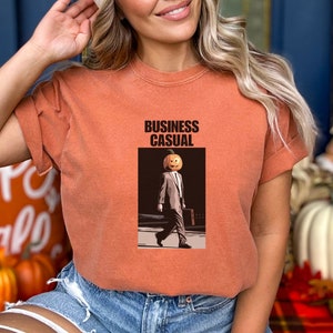 May include: Orange-colored t-shirt with the words "BUSINESS CASUAL" printed at the top. The graphic depicts a person with a pumpkin head wearing a suit and carrying a briefcase. The shirt is a fall-themed design.