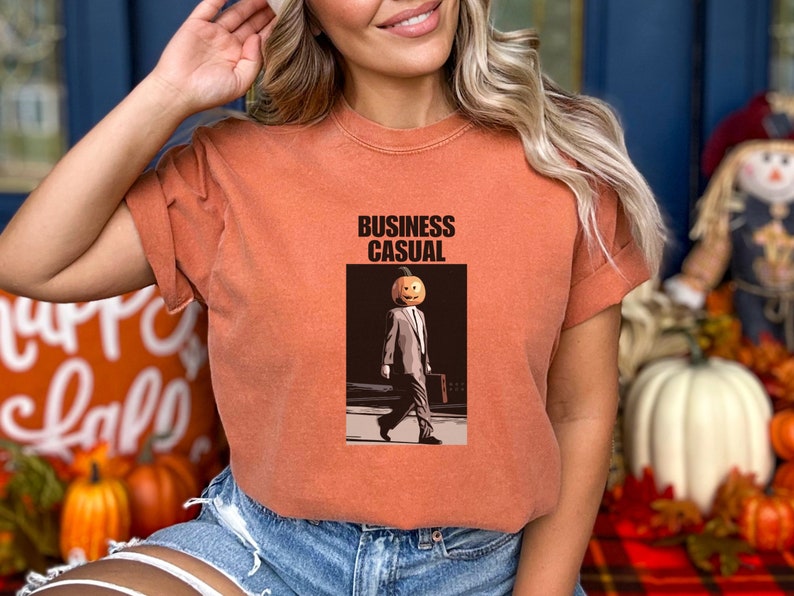 May include: Orange-colored t-shirt with the words "BUSINESS CASUAL" printed at the top. The graphic depicts a person with a pumpkin head wearing a suit and carrying a briefcase. The shirt is a fall-themed design.