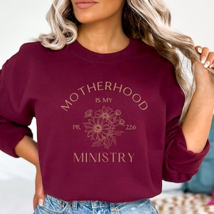 Puede incluir: Sudadera color burdeos con el texto "Motherhood is my ministry" en letras doradas con un dise&ntilde;o floral y el texto "PR. 22:6".