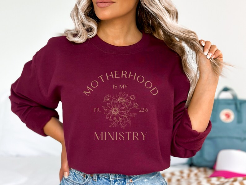 Puede incluir: Sudadera color burdeos con el texto "Motherhood is my ministry" en letras doradas con un dise&ntilde;o floral y el texto "PR. 22:6".