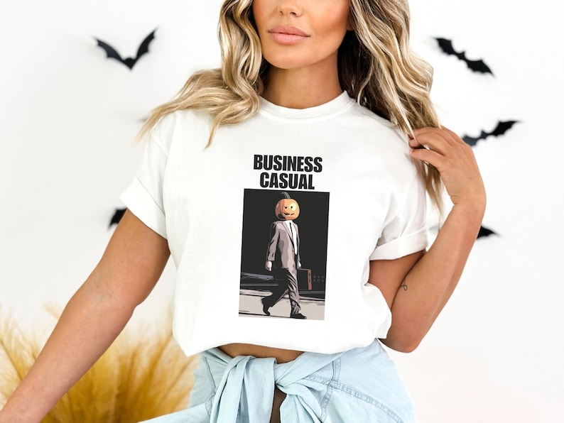 May include: White t-shirt with the words "BUSINESS CASUAL" above an illustration of a person in a suit with a pumpkin head. The person is carrying a briefcase and walking on a crosswalk.