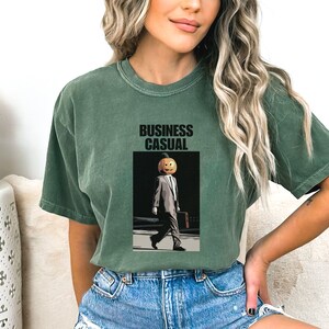 May include: Green t-shirt with the words "BUSINESS CASUAL" above an illustration of a person in a suit with a pumpkin head. The person is carrying a briefcase and walking on a sidewalk.