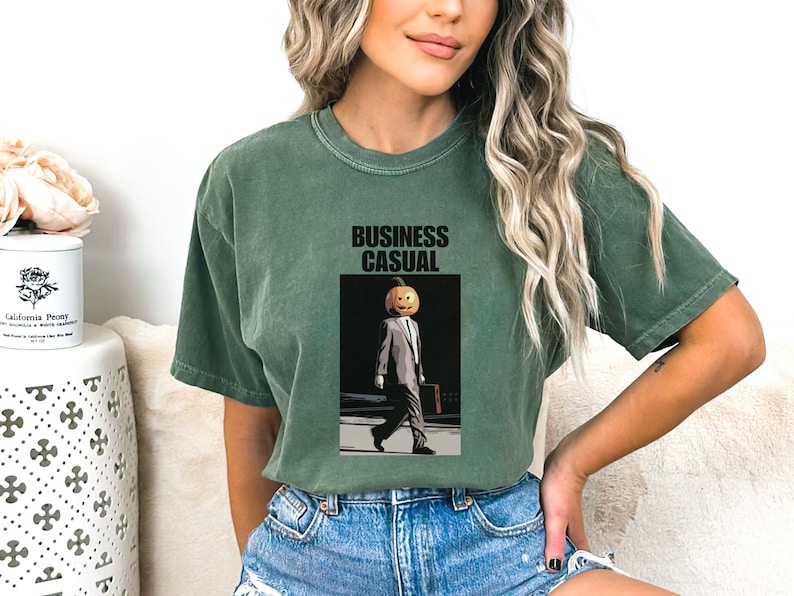 May include: Green t-shirt with the words "BUSINESS CASUAL" above an illustration of a person in a suit with a pumpkin head. The person is carrying a briefcase and walking on a sidewalk.