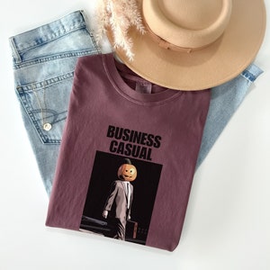 May include: A maroon t-shirt with the words "BUSINESS CASUAL" above an illustration of a person with a pumpkin head wearing a suit. Light blue jeans and a tan hat are also in the image.