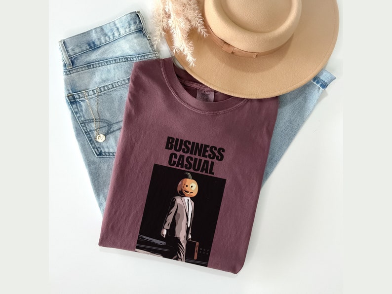 May include: A maroon t-shirt with the words "BUSINESS CASUAL" above an illustration of a person with a pumpkin head wearing a suit. Light blue jeans and a tan hat are also in the image.