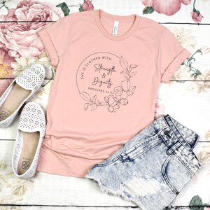 May include: A pink t-shirt with a floral wreath design and the text "She is clothed with Strength & Dignity Proverbs 31:25".
