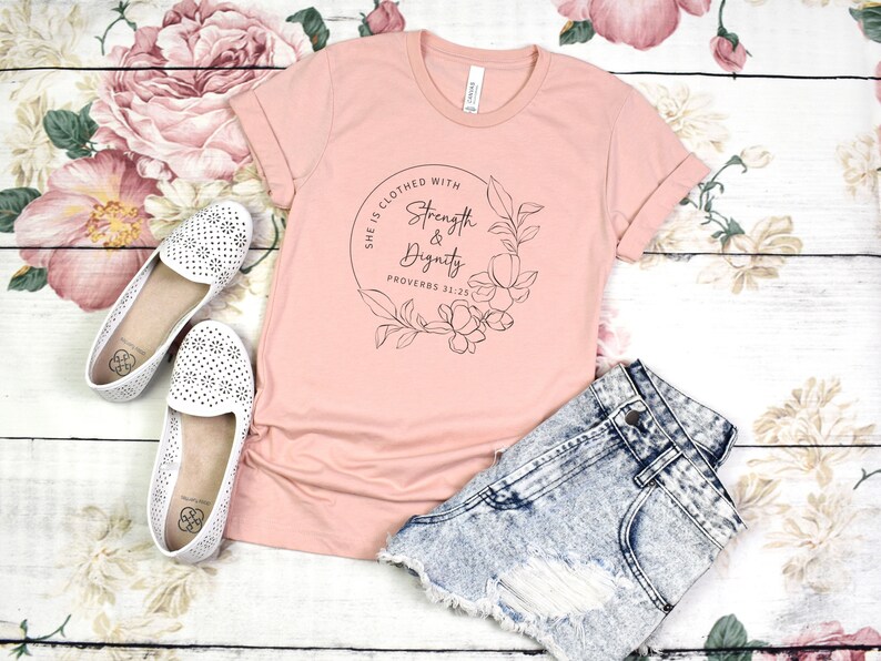 May include: A pink t-shirt with a floral wreath design and the text "She is clothed with Strength & Dignity Proverbs 31:25".