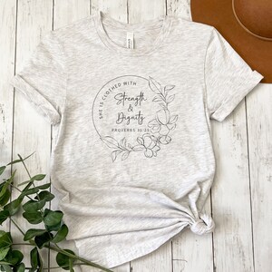 May include: A heather grey t-shirt with a floral design and the text "She is clothed with strength & dignity Proverbs 31:25"