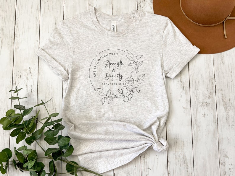 May include: A heather grey t-shirt with a floral design and the text "She is clothed with strength & dignity Proverbs 31:25"