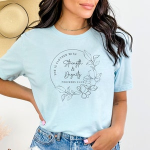 May include: A light blue t-shirt with a black circle design featuring the text "She is clothed with Strength & Dignity Proverbs 31:25" and a floral illustration.