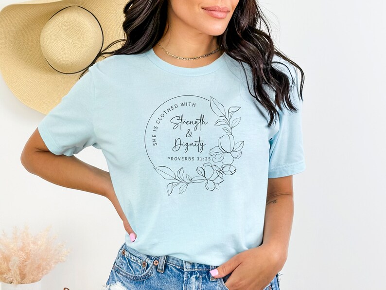 May include: A light blue t-shirt with a black circle design featuring the text "She is clothed with Strength & Dignity Proverbs 31:25" and a floral illustration.