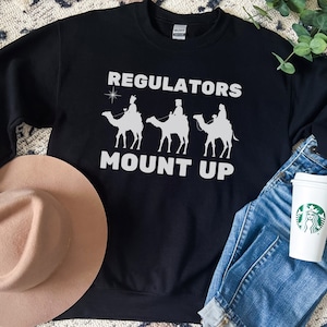 May include: Black sweatshirt with the text "REGULATORS MOUNT UP" in white, featuring a graphic of three camels with riders. A tan hat and a pair of blue jeans are next to the sweatshirt. A white coffee cup with a green logo is also in the image.