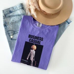 May include: A lavender t-shirt with the words "BUSINESS CASUAL" printed in black. The shirt features a graphic of a person with a pumpkin head wearing a suit and carrying a briefcase. Light blue jeans and a tan hat are also in the image.