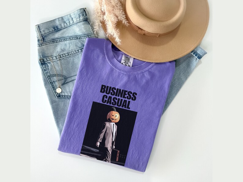 May include: A lavender t-shirt with the words "BUSINESS CASUAL" printed in black. The shirt features a graphic of a person with a pumpkin head wearing a suit and carrying a briefcase. Light blue jeans and a tan hat are also in the image.