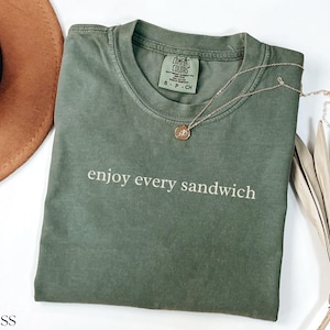 May include: A moss green t-shirt with the phrase "enjoy every sandwich" printed in white. A gold necklace with a pendant rests on the shirt. A brown hat and dried plant are in the background.