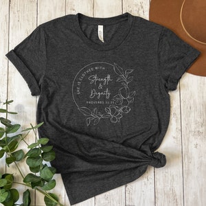 May include: A dark heather grey t-shirt with a white graphic design featuring a floral wreath and the text "She is clothed with strength & dignity Proverbs 31:25".