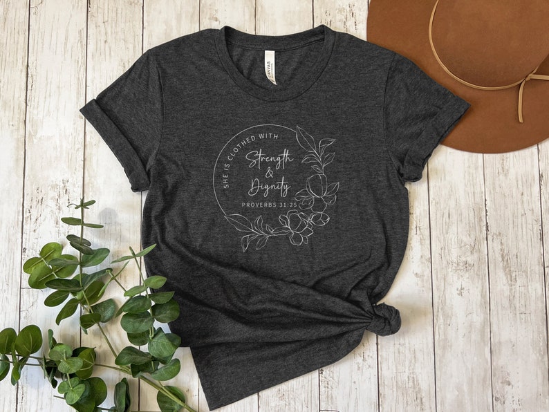 May include: A dark heather grey t-shirt with a white graphic design featuring a floral wreath and the text "She is clothed with strength & dignity Proverbs 31:25".