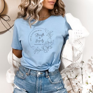 May include: A light blue t-shirt with a black floral design and the text "She is clothed with Strength & Dignity Proverbs 31:25".