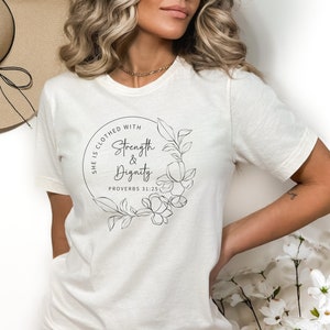 May include: White t-shirt with a black graphic design featuring a floral wreath and the text "She is clothed with Strength & Dignity Proverbs 31:25".