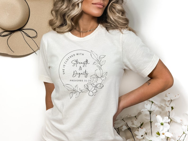 May include: White t-shirt with a black graphic design featuring a floral wreath and the text "She is clothed with Strength & Dignity Proverbs 31:25".