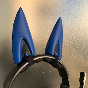 May include: A black headset with bright blue cat ears attached. The ears are secured with a black velcro strap. The headset also features a microphone, perfect for gaming or online communication.