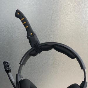 Könnte beinhalten: Schwarzes Gaming-Headset mit einem schwarzen und goldenen Mikrofonarm und einem schwarzen Kopfbügel mit schwarzer Polsterung. Das Headset hat einen schwarzen und goldenen verstellbaren Kopfbügel.