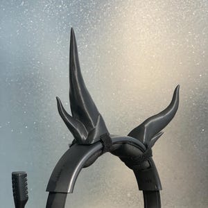 Puede incluir: Auriculares de juego negros con un par de cuernos impresos en 3D unidos a la diadema. Los cuernos son negros y tienen un diseño afilado y puntiagudo.