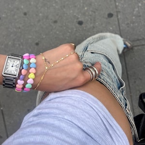 May include: A silver wristwatch with a white face and black Roman numerals is paired with a stack of pastel heart-shaped bead bracelets. A gold hand chain and multiple silver rings complete the jewelry ensemble. Light wash denim jeans are also visible.
