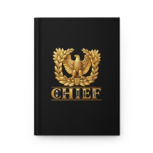 May include: A black hardcover journal featuring a centered gold emblem of an eagle perched within a laurel wreath. Below the emblem, the word CHIEF is printed in bold, gold, serif capital letters, underlined by a thin gold horizontal line.