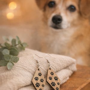 May include: Wooden diamond-shaped earrings with black paw prints. The earrings hang from silver hooks and rest on a folded beige cloth. A dog is blurred in the background.